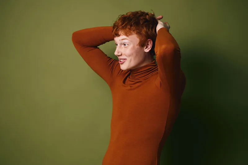 Young man with red hair wearing brown turtleneck sweater poses with hands behind head smiling on green background, casual and confident expression in studio portrait.