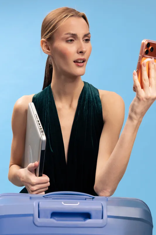 Young woman with laptop and suitcase looks at smartphone with surprised expression on blue background. Concept of travel, technology, business, and modern lifestyle.