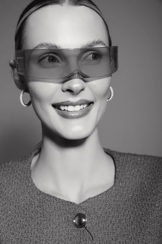Smiling Caucasian woman with fair skin wearing large transparent glasses and pearl earrings. Close-up portrait with smooth hairstyle and textured jacket in black and white style.