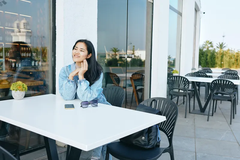 Thinking woman at an outdoor cafe table, resting chin on hands, with smartphone, sunglasses and backpack nearby, daylight, casual mood and relaxed urban moment