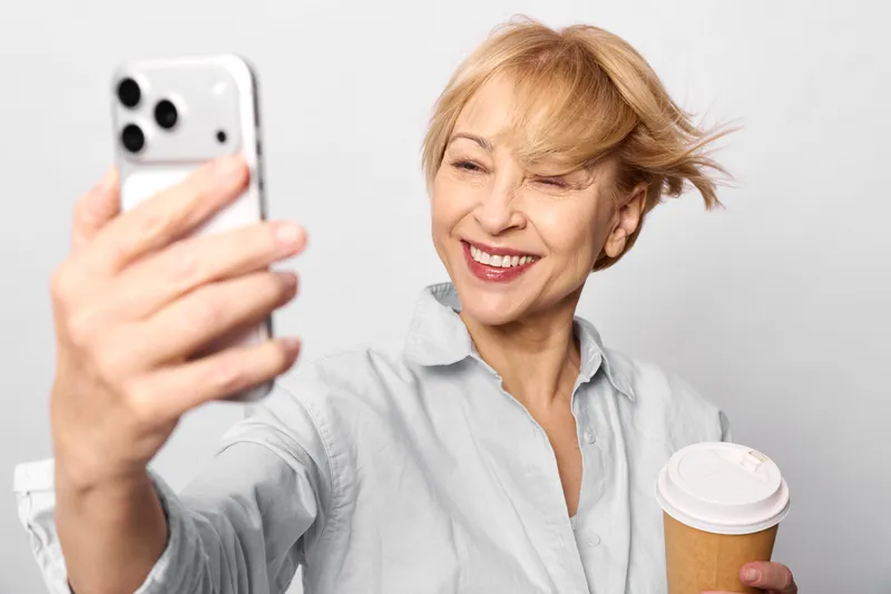 Woman senior smartphone selfie coffee smiling technology communication in a bright studio, she laughs with a paper coffee cup, taking a selfie on a handheld phone