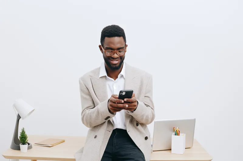 Smiling man using smartphone with laptop and desk accessories enjoying work or leisure in a modern minimalistic workspace. Caucasian, young adult, casual outfit, positive expression, studio shot, clea