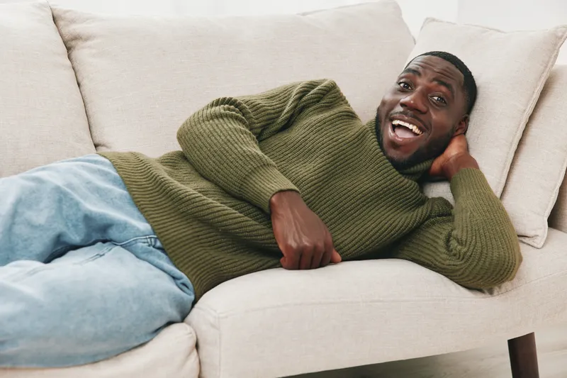 Happy young African man relaxing on a beige sofa, wearing a green sweater and blue jeans, exuding joy and comfort in a serene indoor setting