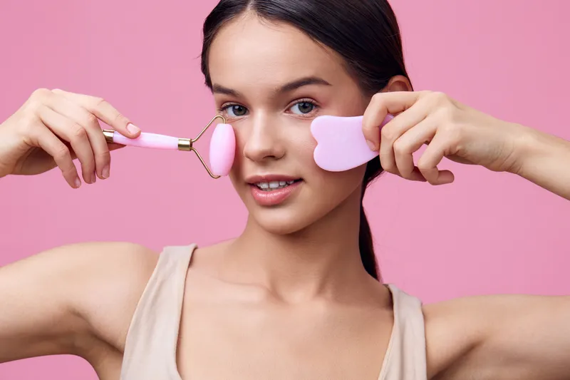 Young woman using facial massage tools on a pink background, promoting skincare and beauty routines with a joyful expression