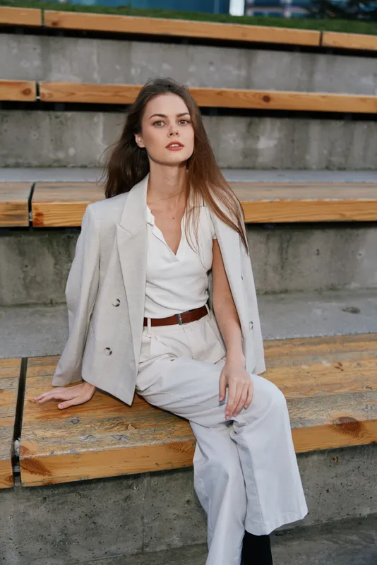 Woman in beige business casual outfit sitting on wooden stairs outdoors, confident young European woman with long brown hair, modern urban fashion style