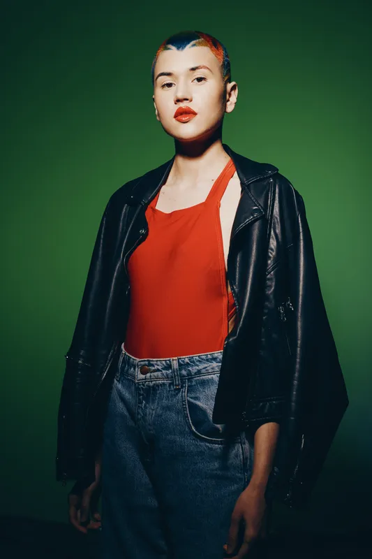 young woman with short colorful hair wearing red top and black leather jacket standing against green background, confident pose, contemporary style