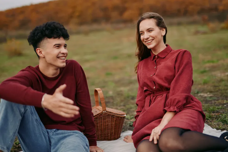 Two friends share an authentic moment on a fall meadow, smiling and talking beside a wicker basket, conveying warmth, calm, and natural connection.