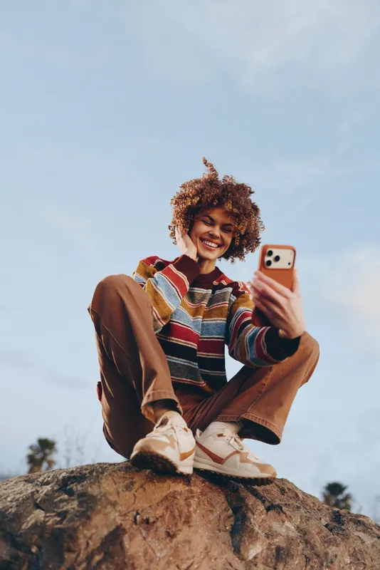Woman lifestyle, curly hair woman sitting on a rock outdoors, smiling at smartphone selfie in warm sunlight, casual sweater, relaxed travel mood, blue sky, distant trees, day trip
