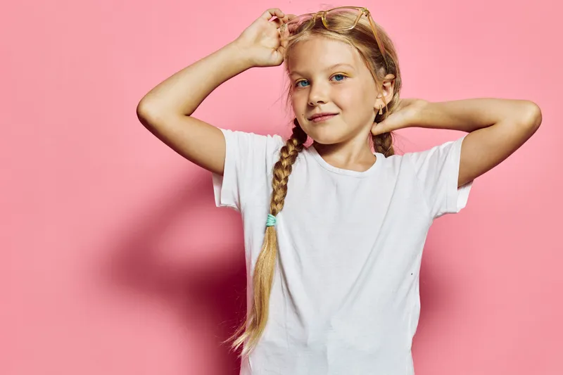 Blonde girl in a white t-shirt poses confidently with braided hair against a pink background. Studio portrait of a child expressing self-assurance and calmness.
