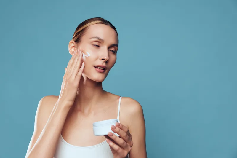 Woman applying skincare cream on face, holding moisturizer in studio, beauty routine for healthy skin, soft light on blue background, calm expression, close up, self care, morning glow