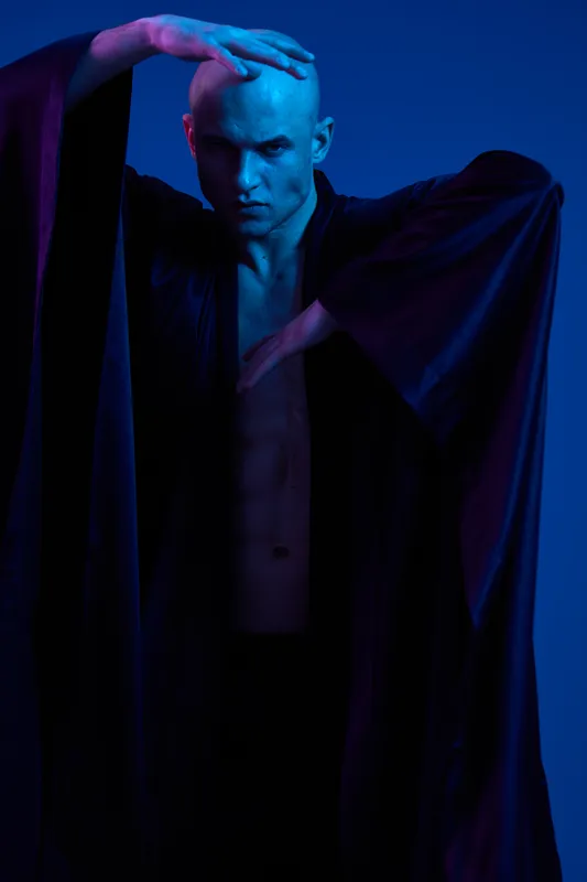 Striking man in black robe poses dramatically against a blue background, showcasing shadows and intriguing lighting, creating a captivating black and white image.