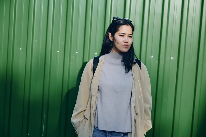 Woman standing beside a green metal wall in a beige coat and gray turtleneck, casual street style, urban vibe, confident look, fashion editorial shot.