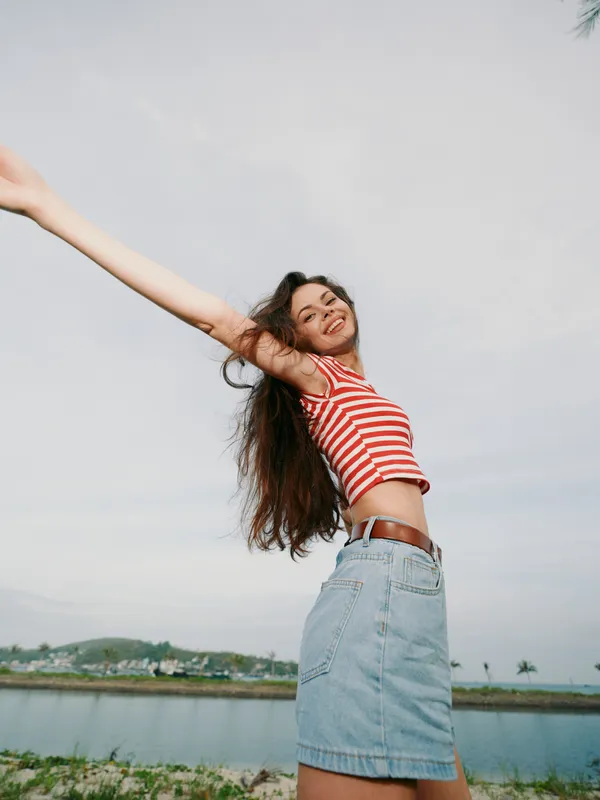 A carefree woman stretches her arms in a bright outdoor scene, wearing a striped red and white crop top and a denim skirt, capturing joyful summer energy.