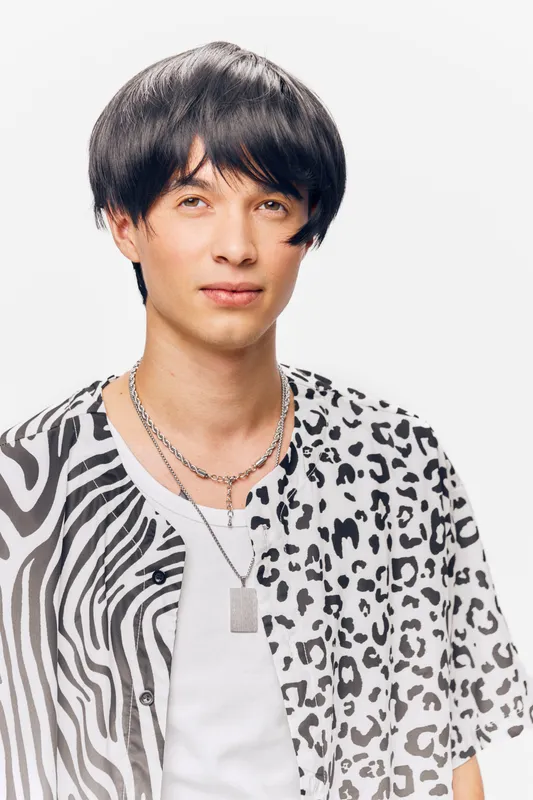 young man wearing patterned black and white shirt with layered necklaces standing on white background with neutral expression, contemporary style portrait