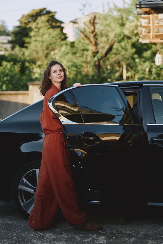 A woman in a red dress stands beside a sleek car, preparing to enter, enjoying a relaxed lifestyle moment outdoors, urban setting, daytime, confident mood