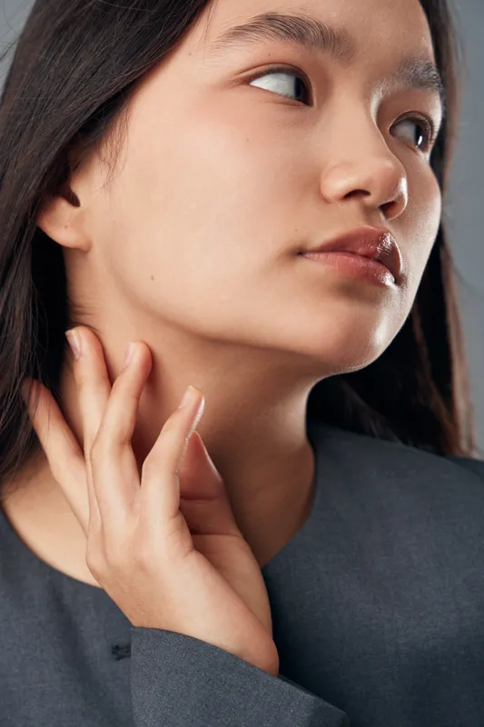 Young woman with clear skin and dark hair touching her neck in closeup portrait with natural makeup and thoughtful expression on neutral background wearing gray outfit
