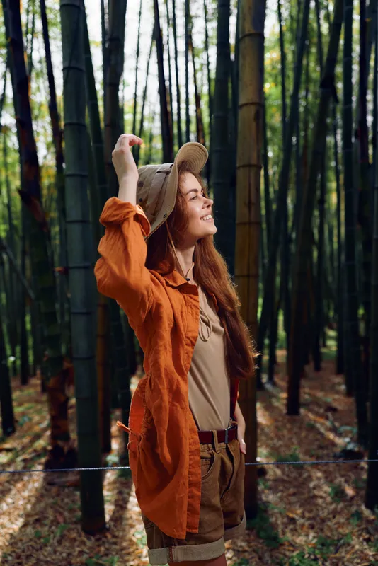 Woman in bamboo forest wearing a hat and orange cardigan, smiling as sunlight filters through trees. Nature portrait of travel, relaxation and peaceful outdoor lifestyle.