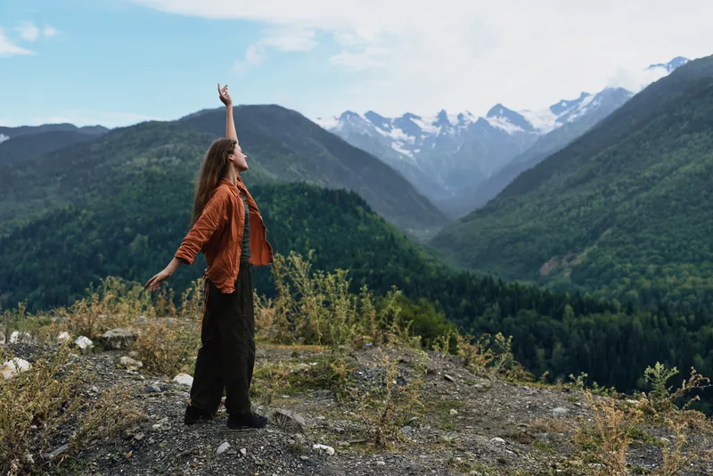 Cheerful traveler standing on a rocky overlook in the mountains, arms raised, enjoying the vast valley and blue sky, embodying freedom, adventure, and a connection with nature