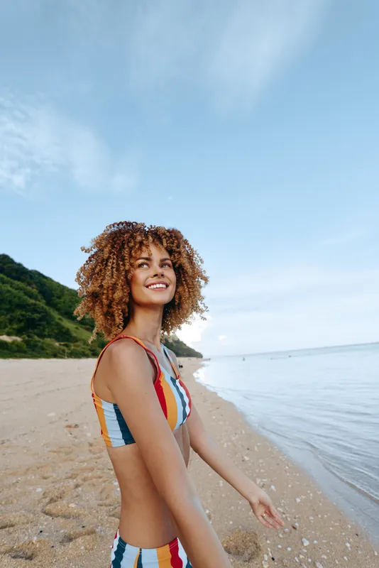 A young woman with curly hair in a striped swimsuit smiles along the beach, standing on warm sand with a calm ocean and a clear blue sky behind.