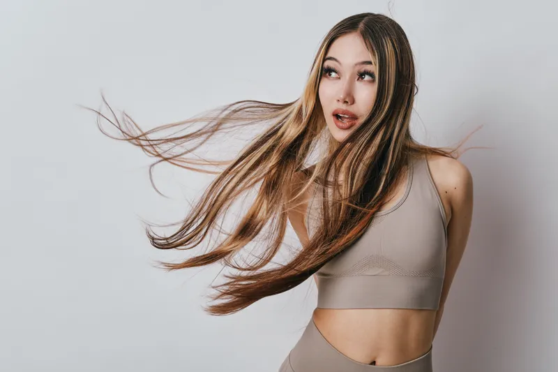 Young woman with long hair wearing sportswear is posing with hair flying and looking sideways on neutral background. Fitness, beauty and motion with active lifestyle model.