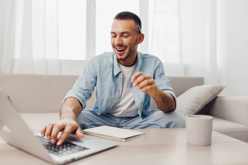 A man smiling while working on his laptop, reflecting themes of soft masculinity and the blend of work-life balance in a modern home.