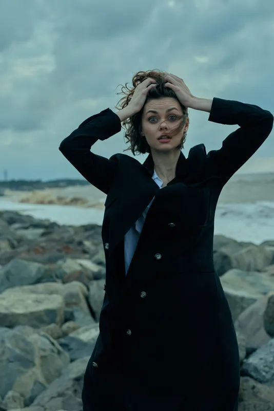 Dramatic wind blown fashion scene with a dark coat at a rocky shoreline, model raises hands to hair, moody skies, ocean spray, dynamic pose, edgy style, outdoor photo shoot with natural elements and
