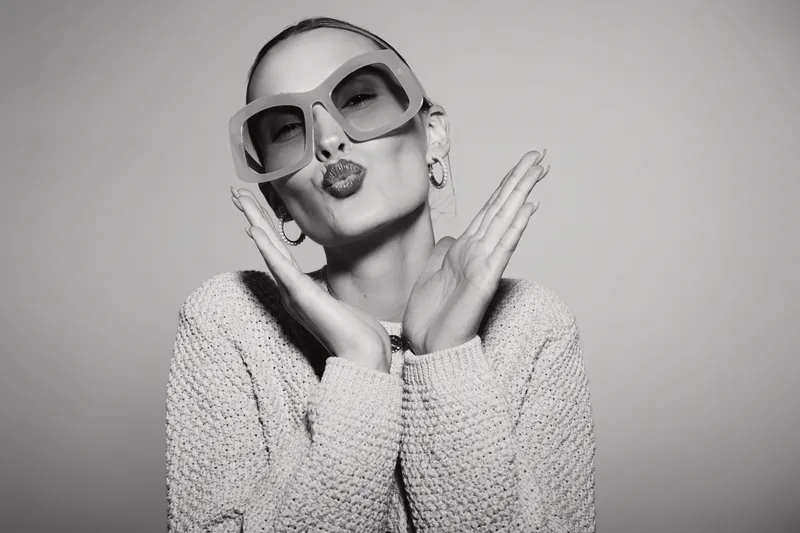 Stylish white woman with pale skin wearing large sunglasses and a knitted sweater blows a kiss. Studio portrait with playful and confident mood.