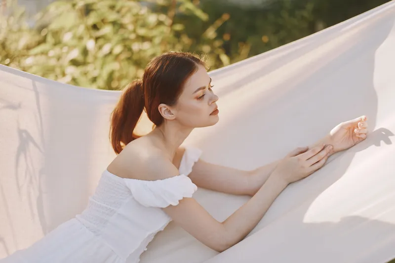 Young woman in a white dress resting on a soft sheet, showcasing serene emotions and a bright outdoor background, conveying tranquility and relaxation in nature
