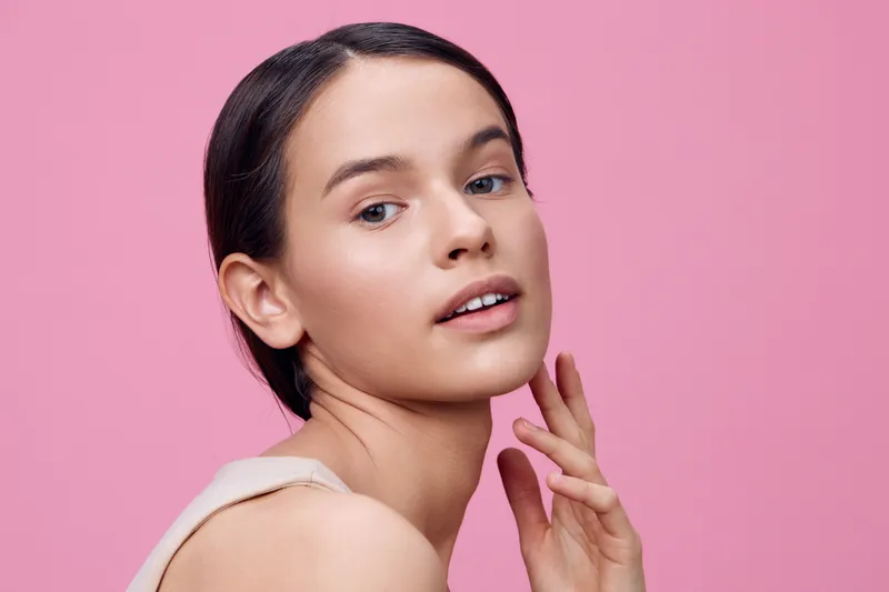 Young model with smooth skin and natural makeup on pink background, conveying beauty, confidence, and elegance in a minimalist style
