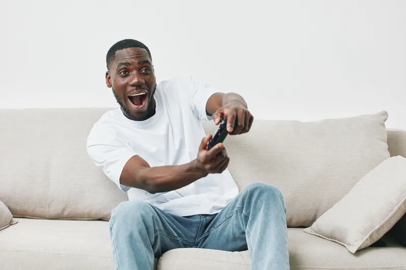 Excited young Black man plays video games on a couch, showcasing joy and engagement in a relaxed home environment with a modern aesthetic