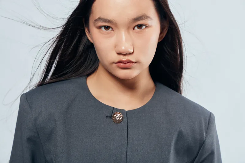 Young woman with long dark hair and serious expression wearing gray jacket standing against light background in studio for fashion portrait or model concept.