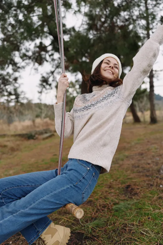 Woman on swing outdoors, joy evident as she wears a cozy sweater and jeans in a park setting, smiling under soft daylight amid autumn trees.