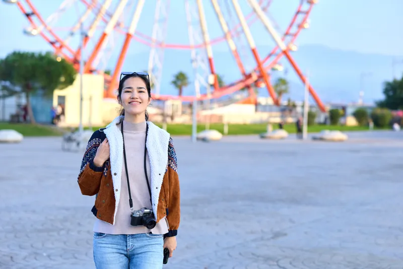 Woman with camera in outdoor park near a ferris wheel, wearing a jacket and scarf, smiling and ready for travel, capturing candid street photography and joyful landscapes in bright daylight for