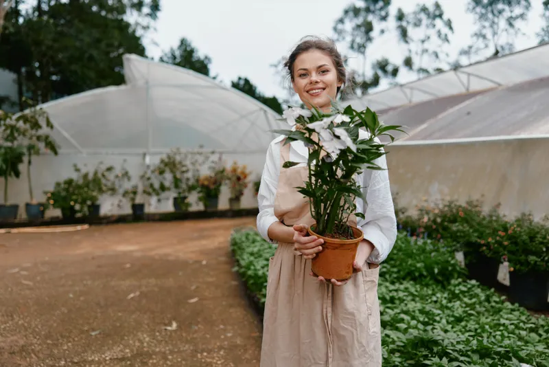 Young woman in a greenhouse holding a potted plant, showcasing a passion for gardening, flourishing greenery, bright colors, and a cheerful atmosphere throughout the scene