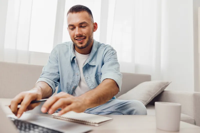 Smiling man working on a laptop, casually dressed in a light blue outfit, surrounded by a bright, modern living space. A cozy atmosphere that enhances focus and productivity.