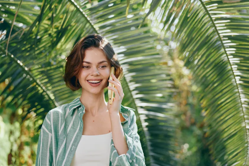 Cheerful woman outdoors talking on phone, smiling warmly amid lush tropical palms and sunlight.