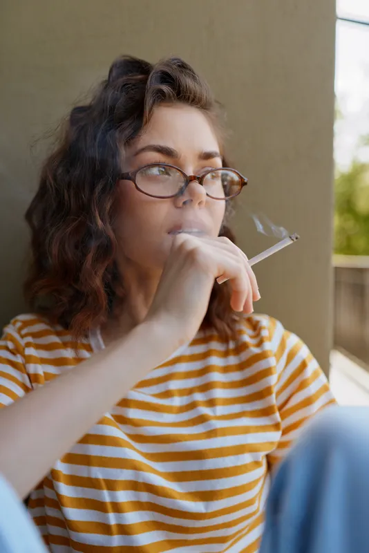 Candid woman with curly hair and glasses smoking a cigarette while seated on a porch, wearing a striped shirt. Warm daylight highlights a relaxed, everyday moment with a calm mood.