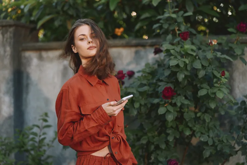 Woman lifestyle portrait outdoors in an orange shirt, standing near blooming roses, holding a smartphone, candid casual mood, relaxed pose in a quiet garden setting