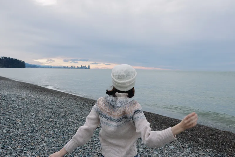 Beach scene with a person facing the sea, arms open, wearing a knit sweater and hat, calm coastline and distant horizon over water.