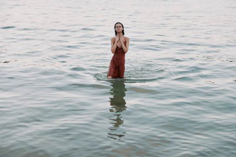 A lone figure stands waist-deep in tranquil sea water, wearing rust-colored overalls, hands pressed near chest, reflecting calm mood and subtle ripples.