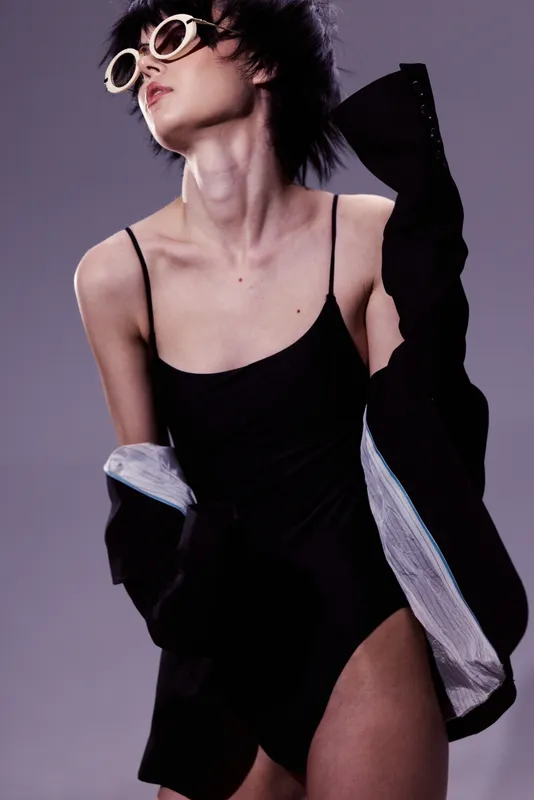 Stylish woman in black bodysuit and sunglasses posing with soft purple background and elegant attitude.
