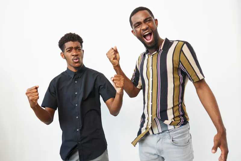 Excited black men expressing joy, celebrating triumph with fists raised, showcasing camaraderie and vibrant energy against a neutral background.
