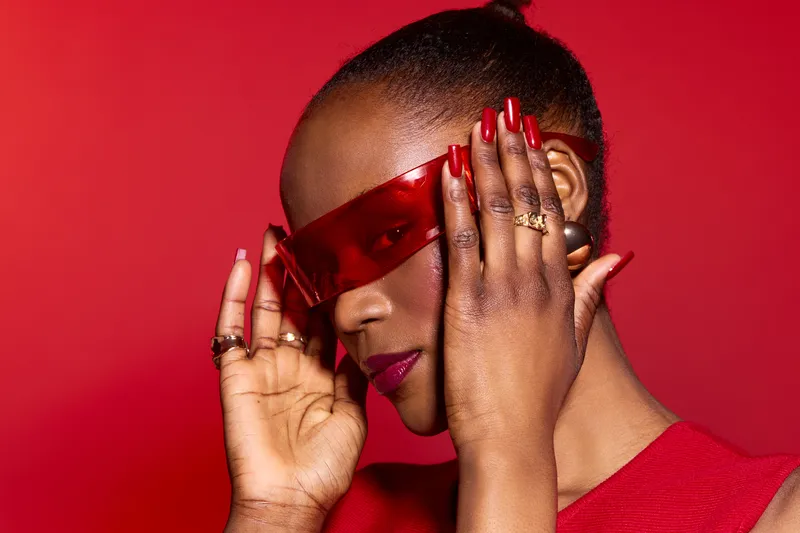 Stylish African woman with dark skin wears red sunglasses and red nails, posing confidently against a red background. Concept of fashion, beauty, and modern style.