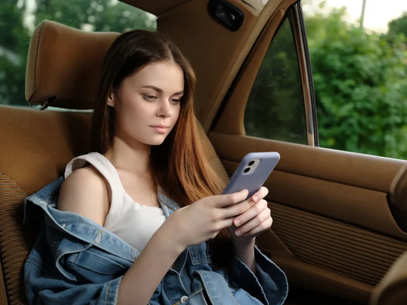 Young woman with long brown hair using smartphone while sitting in a car, wearing a casual outfit, conveying a relaxed and focused emotion, with a natural green background.