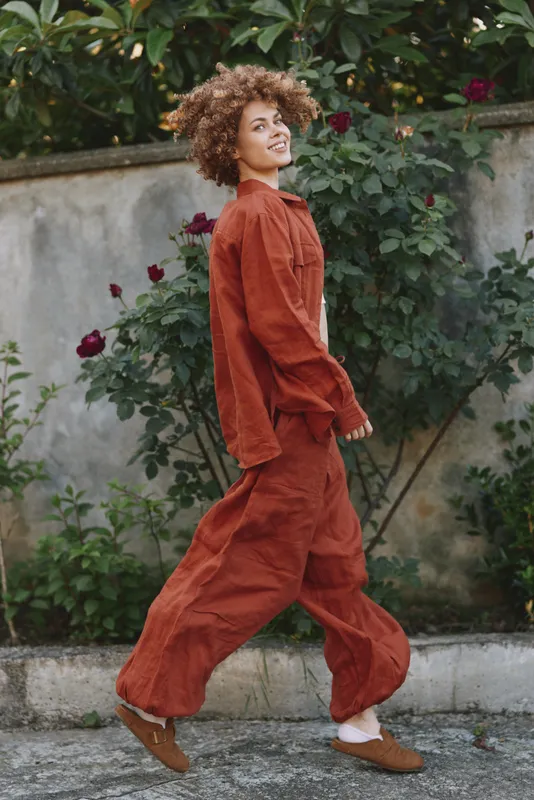 A woman in rust jumpsuit walks along a garden path, smiling with curly hair. Bright outdoor light highlights fabric texture and casual style, embodying everyday confidence