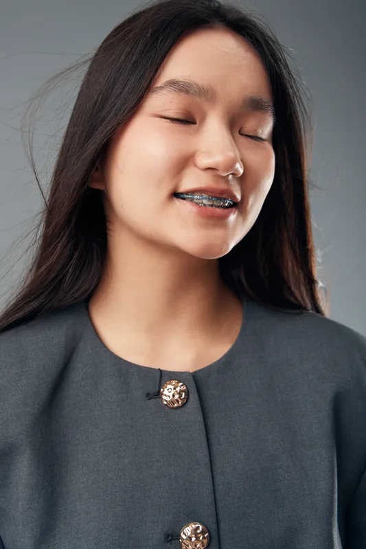 Young woman smiling with closed eyes wearing braces on teeth and long dark hair. Portrait with casual gray outfit and decorative buttons on neutral gray background in studio lighting.