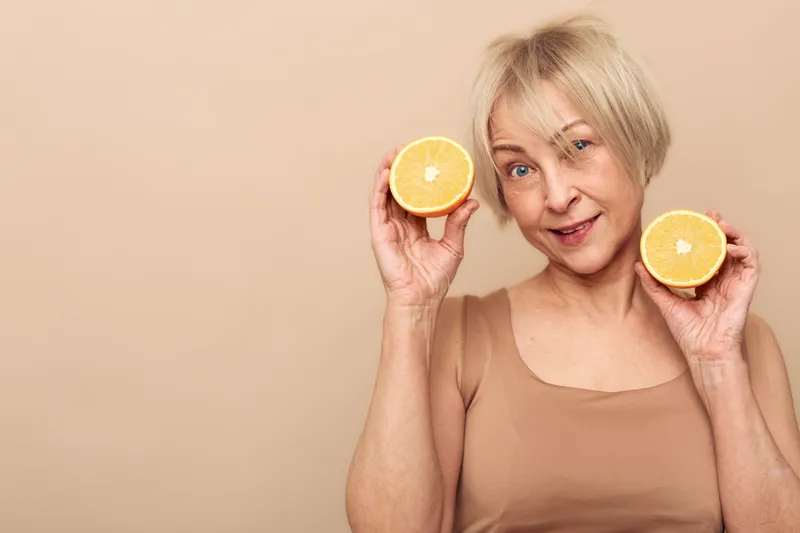 Smiling mature woman with short blonde hair holding two halves of a fresh orange fruit near her face. Concept for healthy lifestyle, natural vitamins, and positive wellness on beige background.