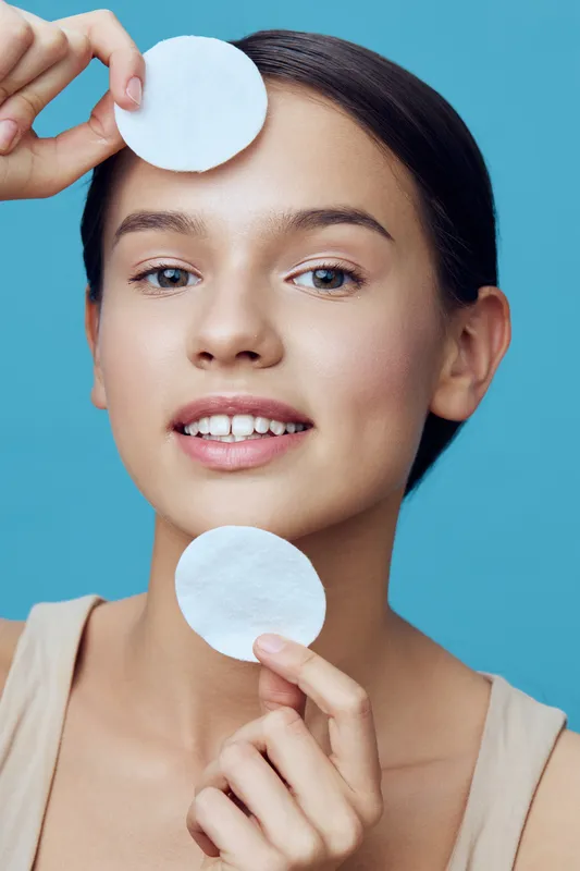 Young woman with natural beauty holding cotton pads, smiling against a turquoise background, representing skincare and freshness