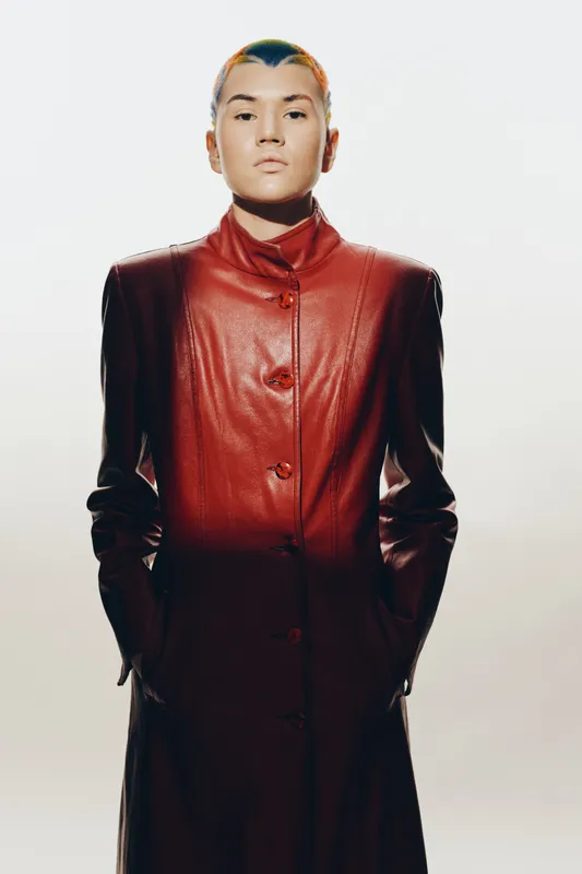 woman in red leather coat standing confidently with hands in pockets and futuristic hairstyle against plain background, showcasing modern fashion.