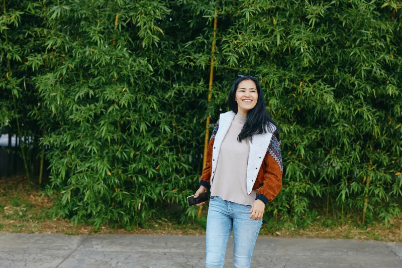 Smiling woman standing outdoors in front of lush greenery, wearing a patterned jacket and light denim jeans, capturing a candid casual lifestyle moment for outdoor photography projects.